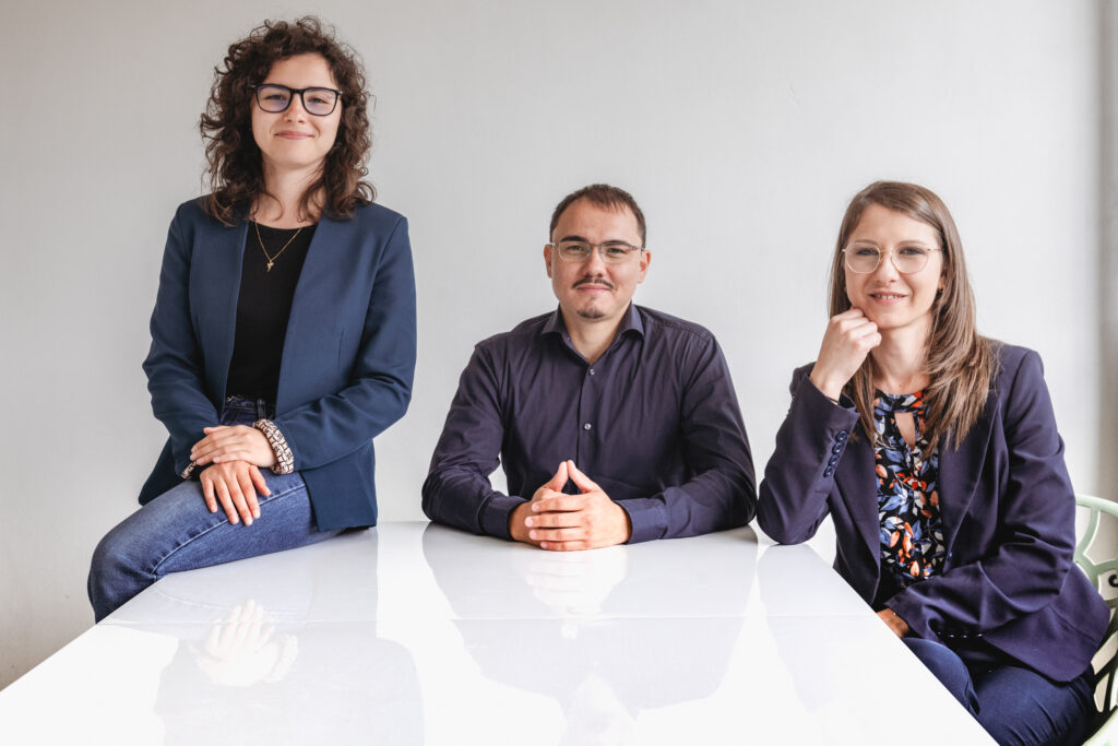 Tre professionisti sorridenti seduti a un tavolo bianco in un ambiente luminoso, vestiti in abiti formali.